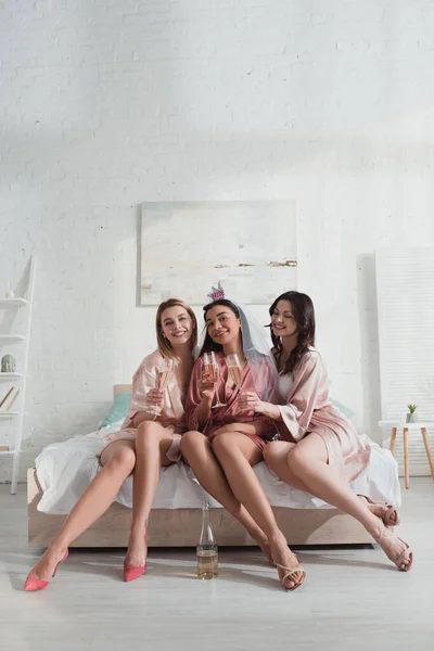 Mujeres multiétnicas felices con copas de champán mirando a la cámara y sonriendo en la cama en la despedida de soltera - foto de stock