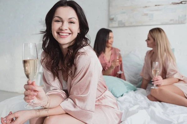 Foco seletivo da menina morena olhando para a câmera, sorrindo e segurando copo de champanhe com amigos multiétnicos na cama — Fotografia de Stock