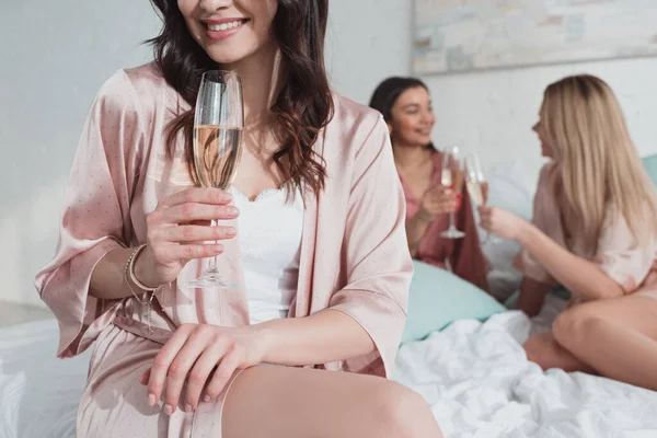 Vista cortada da menina morena sorrindo e segurando copo de champanhe com amigos multiétnicos na cama — Fotografia de Stock