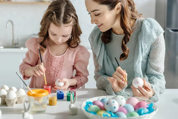 Enfoque selectivo de la madre feliz y la hija pintando huevos de Pascua cerca de conejos decorativos - foto de stock