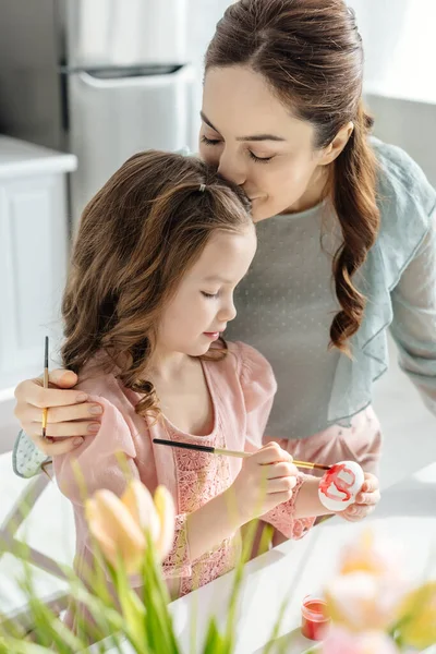 Foyer sélectif de mère embrasser fille mignonne peinture Pâques oeuf près de tulipes — Photo de stock