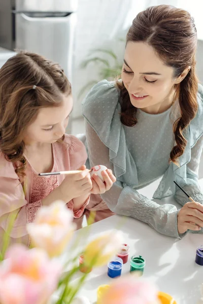 Selektiver Fokus der glücklichen Mutter beim Betrachten der niedlichen Tochter beim Bemalen von Ostereiern bei Tulpen — Stockfoto