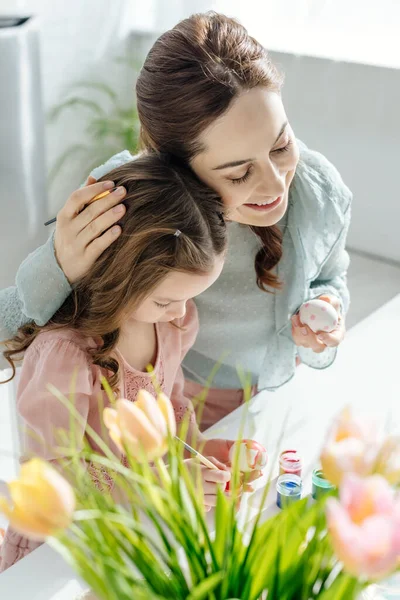 Избирательный фокус счастливой матери, обнимающей милую дочь, рисующую пасхальное яйцо рядом с тюльпанами — стоковое фото