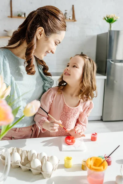 Selective focus of happy kid and mother looking at each other near tulips and chicken eggs — Stock Photo