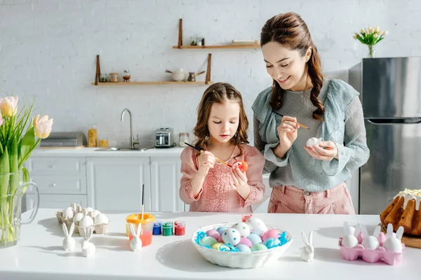 Bambino carino e felice madre pittura uova di pollo vicino conigli decorativi, pane pasquale e tulipani — Foto stock