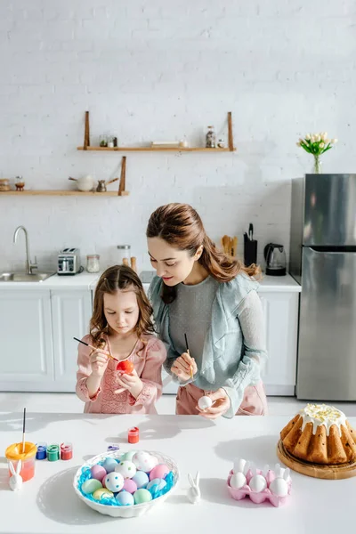 Cute child and happy mother painting easter eggs near decorative rabbits and easter bread — Stock Photo