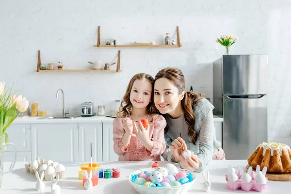 Adorable enfant et mère heureuse près des œufs de poulet peints, lapins décoratifs, pain de Pâques et tulipes — Photo de stock