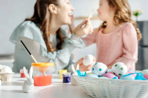 Foco seletivo de ovos de páscoa pintados perto de coelhos decorativos, mãe feliz e filha — Fotografia de Stock