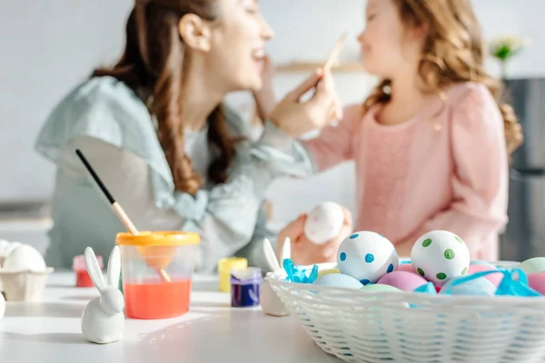 Foco seletivo de ovos de páscoa pintados perto de coelhos decorativos, mãe feliz e filha — Fotografia de Stock