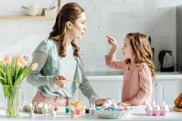 Side view of cheerful mother and happy daughter looking at each other near painted easter eggs and flowers — Stock Photo