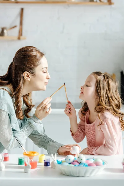 Vista lateral da mãe alegre e filha feliz segurando pincéis perto de ovos de páscoa pintados e coelhos decorativos — Fotografia de Stock