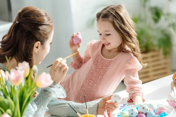 Mãe e criança alegre pintando ovos de Páscoa perto de flores — Fotografia de Stock