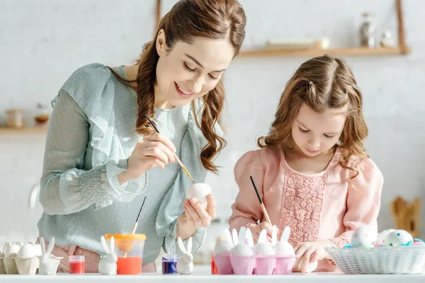 Fröhliche Mutter und glückliches Kind beim Bemalen von Ostereiern neben dekorativen Hasen — Stockfoto