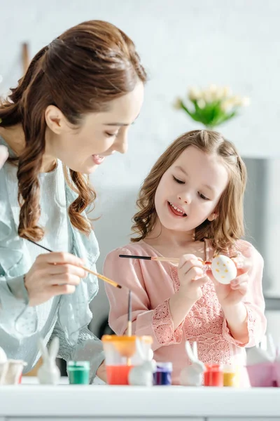 Enfoque selectivo de atractiva madre e hija pintando huevos de Pascua cerca de conejitos decorativos - foto de stock