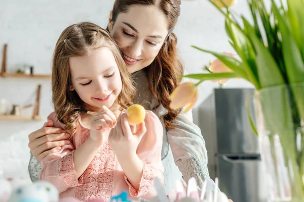 Foco seletivo de tulipas perto de mãe feliz e filha pintando ovo de Páscoa — Fotografia de Stock