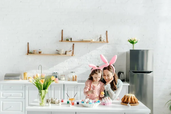 Счастливые ребенок и мать с кроличьими ушами рядом куриные яйца, декоративные кролики, пасхальный хлеб и тюльпаны — стоковое фото
