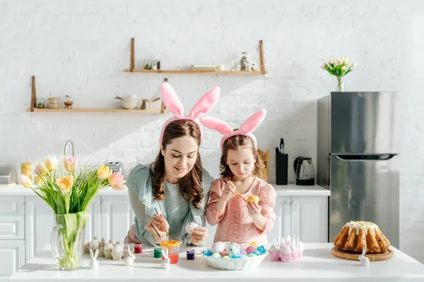 Niño feliz y atractiva madre con orejas de conejo cerca de huevos de pollo, conejos decorativos, pan de Pascua y tulipanes - foto de stock