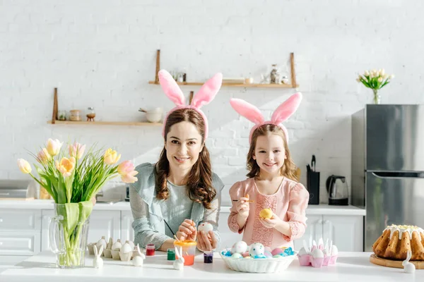 Criança feliz e mãe atraente com orelhas de coelho perto de ovos de Páscoa, coelhos decorativos e tulipas — Fotografia de Stock