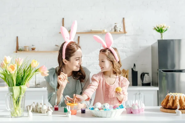 Bambino felice e madre con orecchie di coniglio vicino alle uova di pollo, conigli decorativi, pane pasquale e tulipani — Foto stock