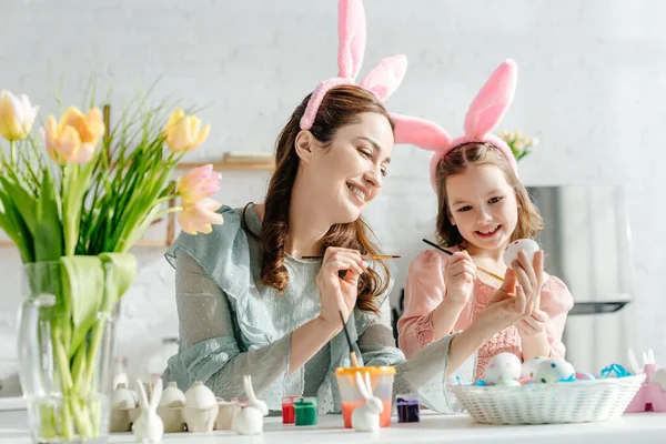 Selektiver Fokus von Mutter und Kind in Hasenohren beim Bemalen von Hühnereiern neben Tulpen — Stockfoto