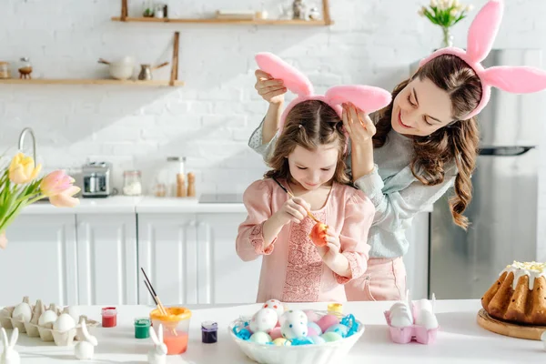 Enfoque selectivo de madre feliz tocando orejas de conejo en hija pintando huevo de gallina cerca de pan de Pascua - foto de stock