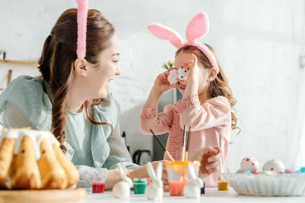 Foco seletivo da mãe feliz olhando para a filha em orelhas de coelho cobrindo olhos com ovos de Páscoa — Fotografia de Stock