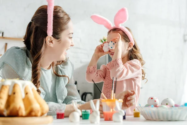 Selective focus of happy mother looking at daughter in bunny ears covering eyes with easter eggs — Stock Photo