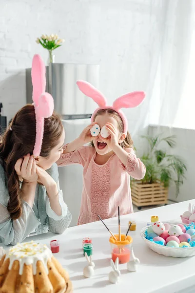 Селективное внимание матери, смотрящей на милую дочь в кроличьих ушах, закрывающих глаза пасхальными яйцами — стоковое фото