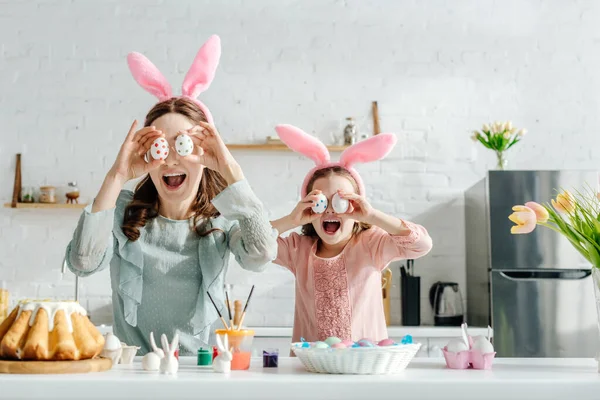 Excitada madre e hija en orejas de conejo cubriendo los ojos con huevos de pollo pintados cerca de tulipanes y pan de Pascua - foto de stock