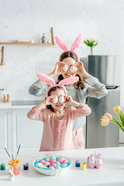 Happy mother and daughter in bunny ears covering eyes with easter eggs near tulips — Stock Photo