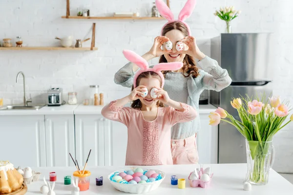 Sonrientes madre e hija en orejas de conejo cubriendo los ojos con huevos de Pascua cerca de tulipanes - foto de stock