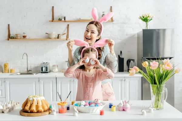 Mãe feliz tocando orelhas de coelho na filha cobrindo olhos com ovos de galinha perto de pão de Páscoa e tulipas — Fotografia de Stock