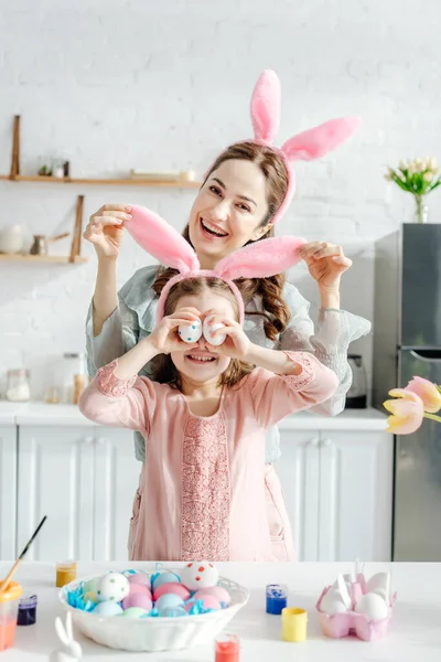 Happy mother touching bunny ears on daughter covering eyes with chicken eggs near tulips — Stock Photo