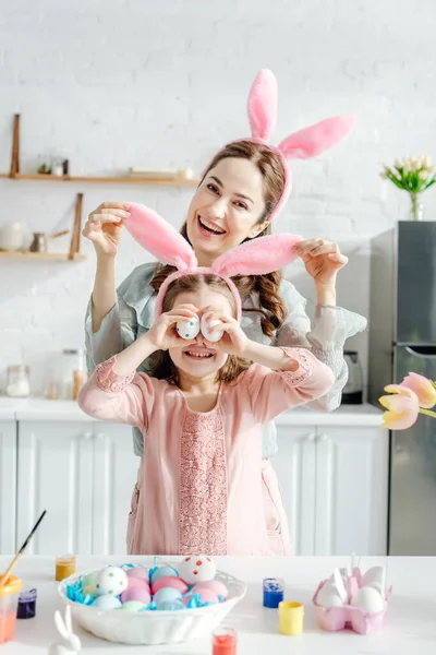 Feliz madre tocando conejito orejas en hija cubriendo ojos con huevos de pollo cerca de tulipanes - foto de stock