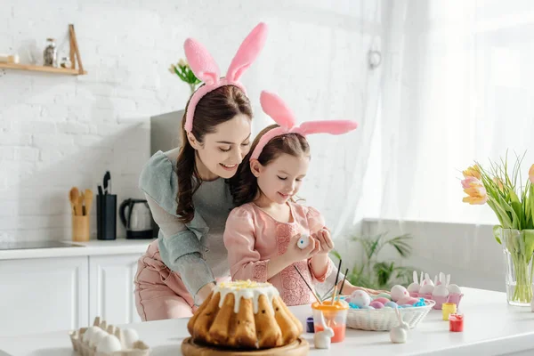 Feliz madre e hija en orejas de conejo mirando huevos de pollo cerca de tulipanes y pan de Pascua - foto de stock