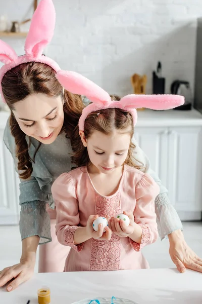 Feliz madre e hija en orejas de conejo mirando huevos de Pascua - foto de stock