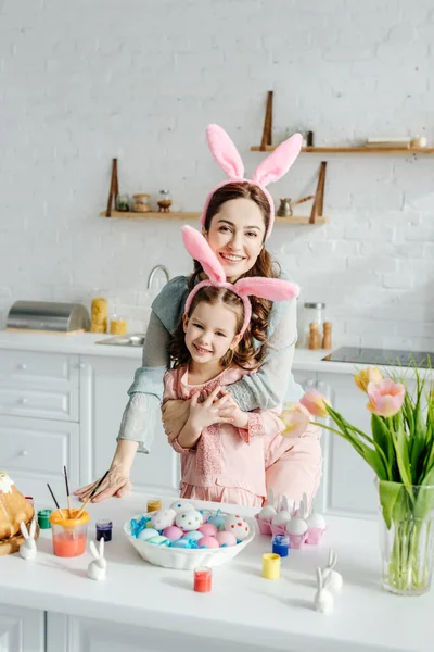 Mère heureuse embrassant fille dans les oreilles de lapin près des oeufs de Pâques — Photo de stock