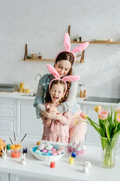 Mère heureuse embrassant fille excitée dans les oreilles de lapin près des oeufs de Pâques — Photo de stock