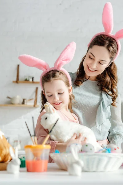 Madre feliz mirando al niño en orejas de conejo con conejo de juguete - foto de stock