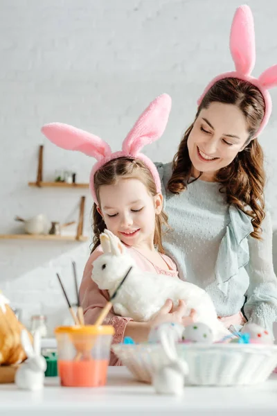 Madre feliz mirando al niño en orejas de conejo con conejo de juguete - foto de stock