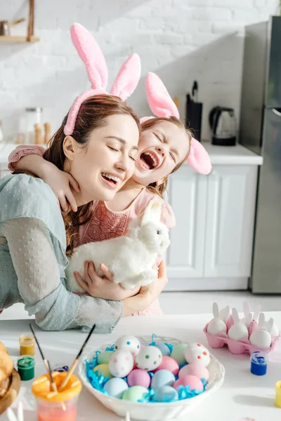 Mãe feliz abraçando filha bonito em orelhas de coelho com coelho de brinquedo — Fotografia de Stock
