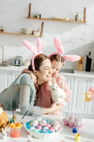 Mãe alegre abraçando filha bonito em orelhas de coelho com coelho de brinquedo — Fotografia de Stock