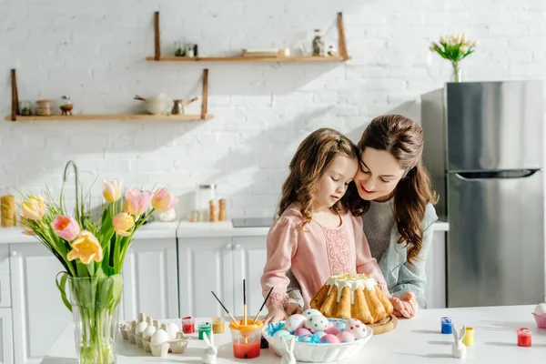 Bambino adorabile e madre felice vicino alle uova di Pasqua, conigli decorativi, pane pasquale e tulipani — Foto stock