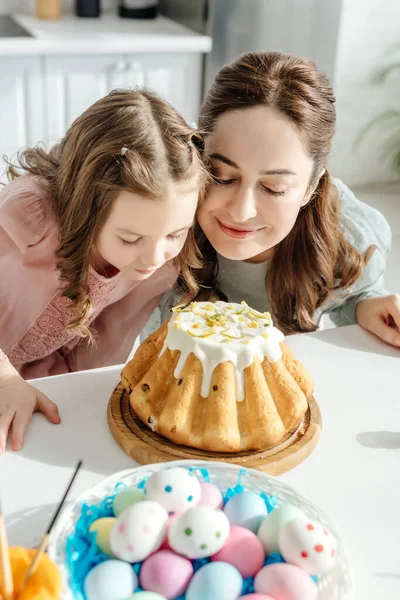 Foco seletivo de feliz mãe e filha cheirando bolo de Páscoa perto de ovos de galinha pintados — Fotografia de Stock