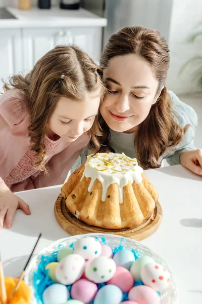 Selektiver Fokus glücklicher Mutter und Tochter, die Osterkuchen neben bemalten Hühnereiern riechen — Stockfoto