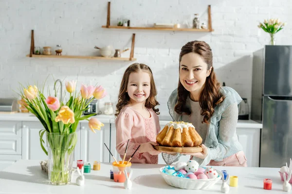 Criança adorável e mãe atraente perto de ovos de Páscoa, coelhos decorativos, pão de Páscoa e tulipas — Fotografia de Stock