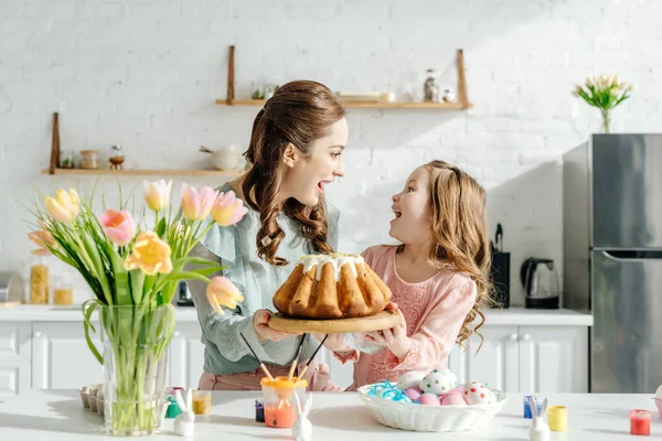 Mãe animada e criança olhando uns para os outros perto de ovos de Páscoa, coelhos decorativos, pão de Páscoa e tulipas — Fotografia de Stock