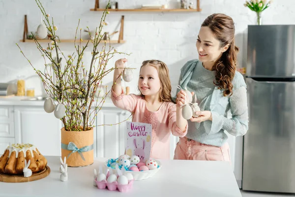 Criança alegre tocando ovo de Páscoa decorativo perto do salgueiro de mãe e coelhos decorativos — Fotografia de Stock