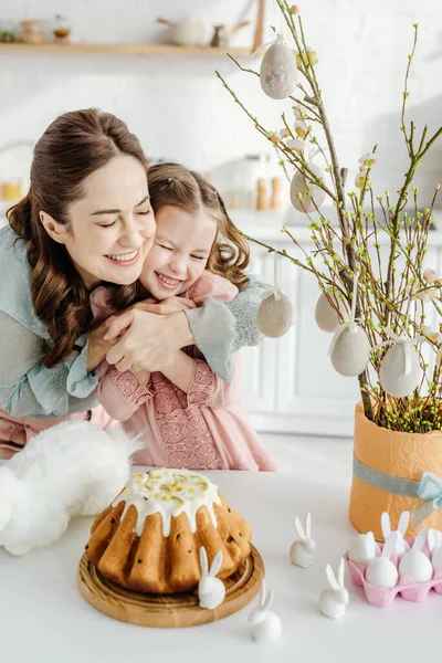 Mãe feliz abraçando filha perto de vaso de flores willowin, coelhos decorativos e bolo de Páscoa — Fotografia de Stock