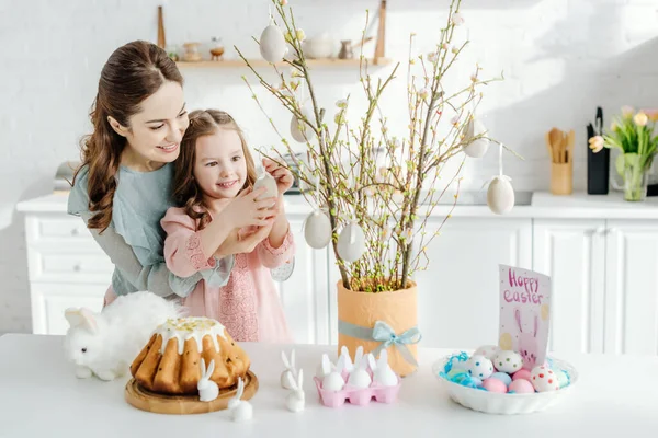 Glückliches Kind berührt dekoratives Osterei auf Weidenzweig in der Nähe der Mutter — Stockfoto